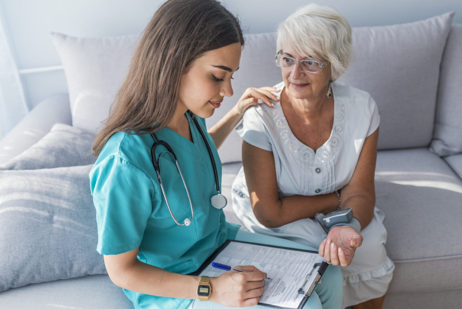 A nurse in teal scrubs talking to an elderly woman on a couch.